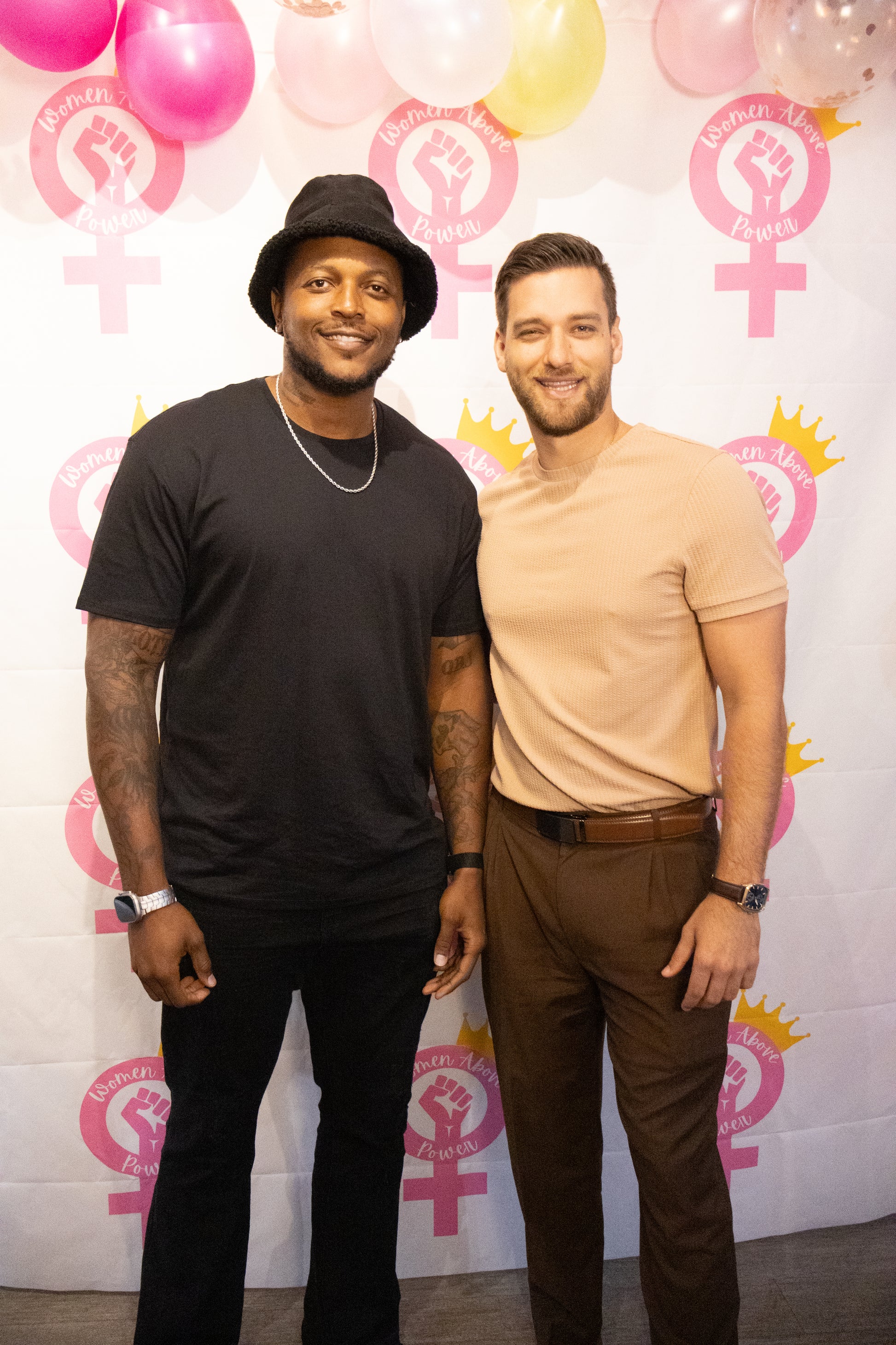 Two men standing in front of a pink and white background with gender symbols and balloons.