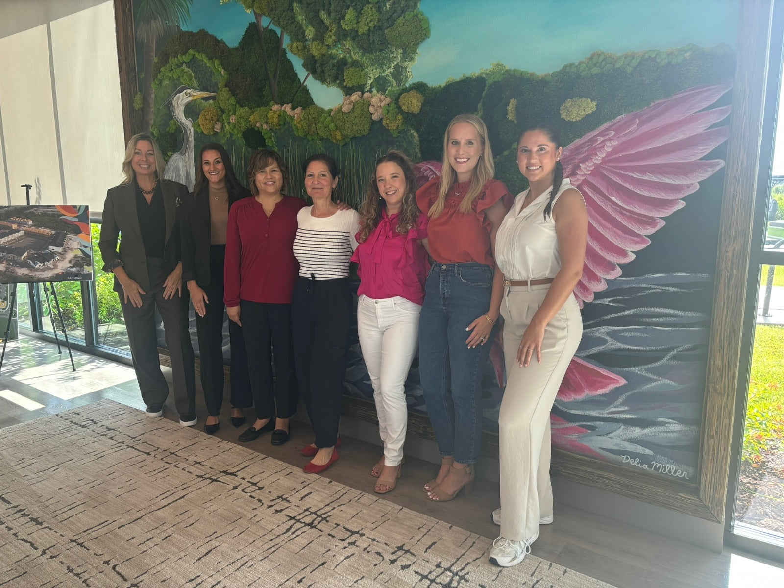 Group of women posing in front of a large painting with pink wings.